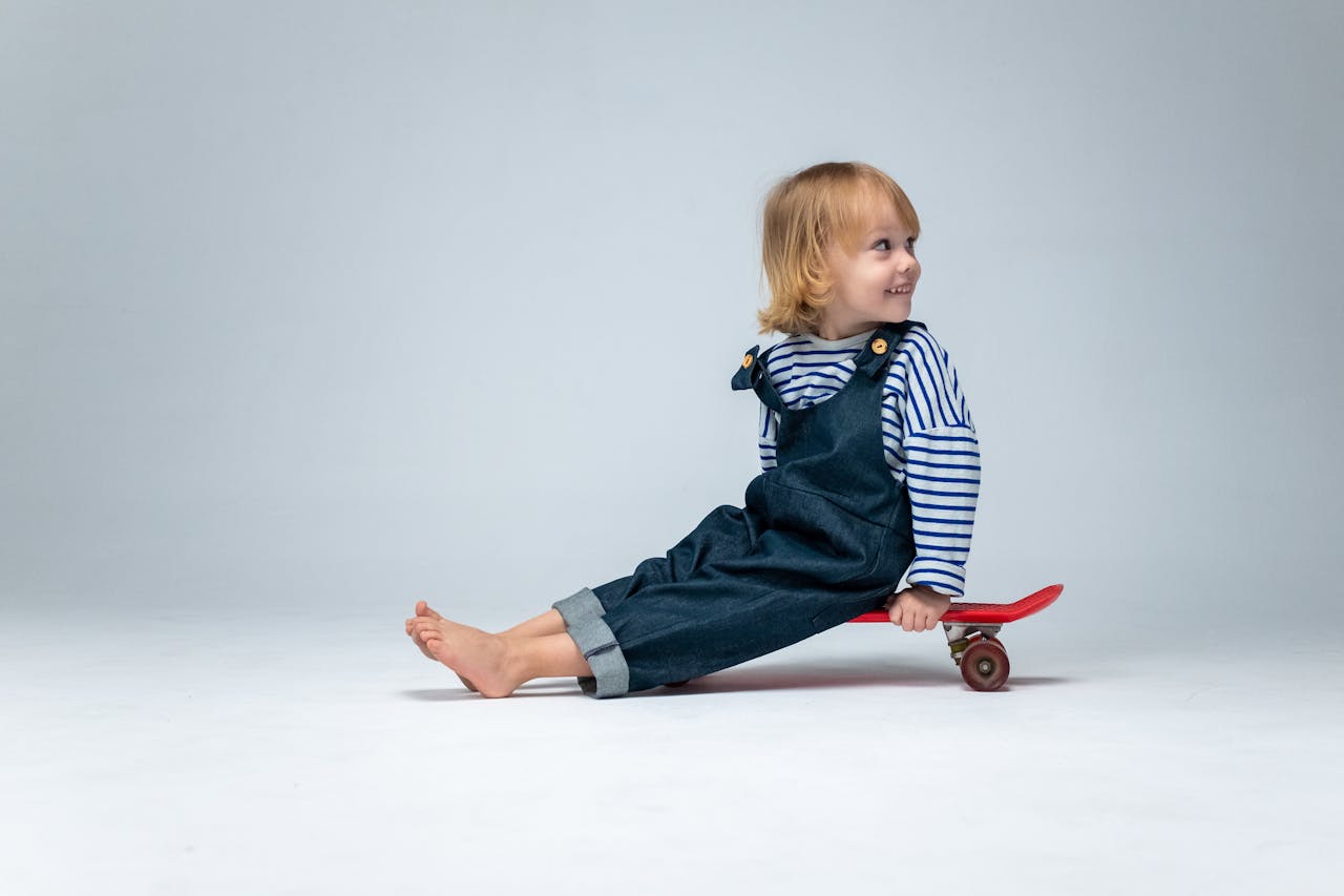 Adorable toddler in striped shirt and overalls on a skateboard in a studio setting, full of joy and playfulness.